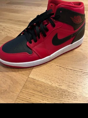 Nike Air Jordan 1 Mid röd/svart - Snygga Nike Air Jordan 1 Mid sneakers i rött och svart med klassisk svart swoosh och Air Jordan-logga på sidan. Skon har svart tåparti, röda paneler och vit mellansula. Yttermaterial i läder och slitstark röd yttersula. Perfekt för dig som gillar streetwear och ikonisk design.