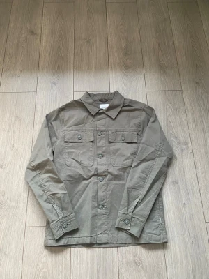 Olivgrön overshirt J.Lindeberg  - Stilren olivgrön overshirt med klassisk krage och knäppning framtill. Två bröstfickor med lock och knappar ger en cool utility-look. Långärmad modell i ett robust bomullstyg, perfekt att bära lager på lager.