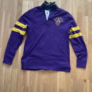 Ralph Lauren piké - Snygg lila pikétröja från Polo Ralph Lauren med gula ränder på ärmarna och broderad logga på bröstet. Tröjan har hög krage med knappar, coola detaljer på armbågarna och stor vit siffra på ryggen. Perfekt för dig som gillar sportig och preppy stil. 