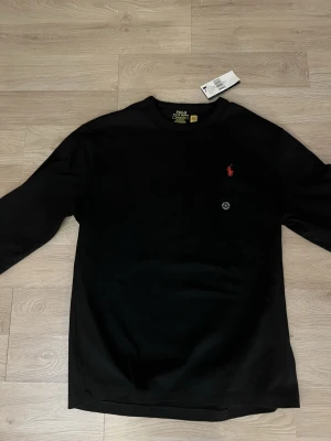 Svart tröja från Polo Ralph Lauren - Svart långärmad tröja från Polo Ralph Lauren i klassisk passform. Tröjan har rund halsringning och den ikoniska röda broderade loggan på bröstet. Perfekt för en stilren och enkel look.