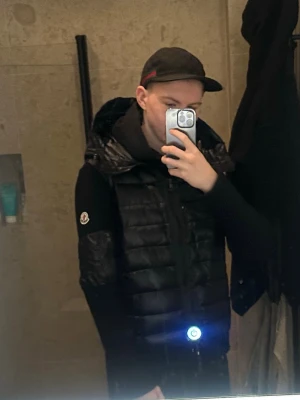 Svart cardigan jacka från Moncler - Svart cardigan jacka från Moncler med quiltad framsida i dun och stickade ärmar. Jackan har huva med snörning, dragkedja framtill och Moncler-logga på ärmen. Perfekt för dig som vill ha en stilren och sportig look.