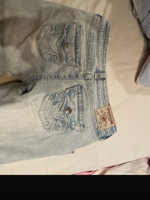 True Religion Joey Low Rise Flare Jeans - Ljusblå jeans från True Religion, modell Joey Low Rise Flare. Klassiska bakfickor med lock och silverfärgade knappar, samt ikonisk patch bak i midjan. Jeansen har låg midja och utsvängda ben för en retro vibe. Aldrig använda modell joey storlek 25 super snygga 