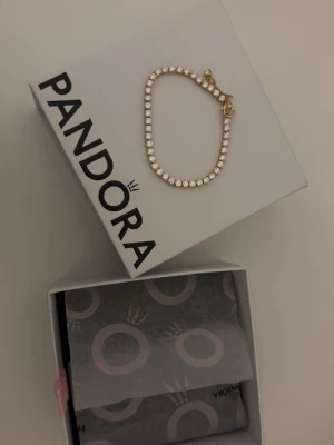 Guldfärgat armband från Pandora - Stilrent armband från Pandora i guldfärg med gnistrande stenar runt hela bandet. Klassisk och elegant design med smidig låsdetalj. Perfekt accessoar för att lyfta din outfit.