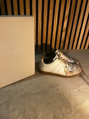 Maison Margiela sneakers med färgstänk - Unika vita sneakers från Maison Margiela med färgstänk i blått, rött, gult och grönt. Skorna har klassisk låg siluett, vita snören och beige sula i gummi. Tillverkade i läder och mocka för en lyxig känsla. Perfekta för dig som vill sticka ut med din stil.