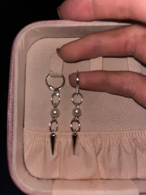 Silvriga örhängen med pärlor och spikes - Säljer ett par snygga örhängen i silverfärg med två små vita pärlor och en spetsig spike-detalj längst ner. Örhängena har en modern design med ringar och hängande detaljer. Perfekta för dig som gillar en edgy stil.