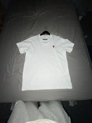 Vit t-shirt med rött hjärta från Ami - Vit t-shirt från Ami med ett litet rött hjärta broderat på bröstet. Klassisk passform och rund halsringning. Perfekt för en clean och stilren look.