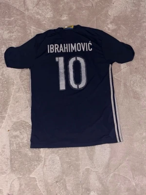 Sverige fotbollströja Ibrahimović #10 - Mörkblå och grå fotbollströja från Adidas med Sveriges landslagsemblem och nummer 10 på framsidan. På ryggen står det Ibrahimović och nummer 10. Tröjan har korta ärmar och klassiska Adidas-ränder på sidorna.