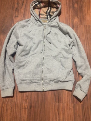 Grå zip-hoodie från Burberry - Snygg grå hoodie med dragkedja från Burberry. Klassisk huva med det ikoniska rutmönstret på insidan och broderad logga på bröstet. Mjuk bomullsblandning, två fickor fram och ribbade muddar. Perfekt för en avslappnad och stilren look.
