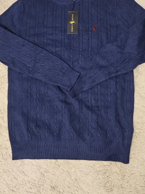 Blå kabelstickad tröja Ralphh Laurennn - Kabelstickad blå tröja från Ralphh Laurenn med rund halsringning och klassisk röd broderad logga på bröstet. Tröjan har ribbade muddar och är perfekt för en stilren look. Materialet känns mjukt och håller formen snyggt.