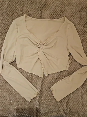 Beige croppad topp från SHEIN - Trendig beige croppad topp från SHEIN med lång ärm och snygg twistad detalj framtill. Tillverkad i mjuk polyesterblandning med elastan för stretchig passform. Toppen har v-ringning och lätt vågig nederkant för en modern look.