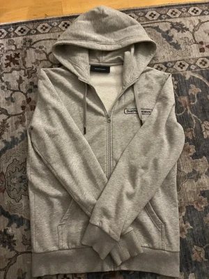 Grå zip-hoodie från Peak Performance - Snygg ljusgrå hoodie med dragkedja från Peak Performance. Klassisk modell med huva och dragsnören, samt fickor framtill. Broderad logga på bröstet. Perfekt för en avslappnad och sportig stil.