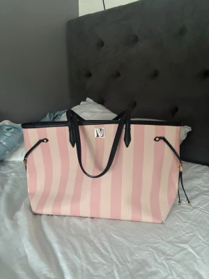 Randig shoppingväska från Victoria Secret 👛 - Säljer en stor shoppingväska från Victoria Secret med rosa och beigea ränder. Väskan har svarta handtag och detaljer samt ett metallfärgat V-logga framtill. Perfekt för dig som vill ha plats för allt och gillar en chic look. Aldrig använd!