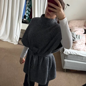 Grå stickad ponchotröja med bälte - Mysig grå stickad ponchotröja med korta ärmar och knytskärp i midjan. Tröjan har hög hals och en grovstickad struktur🩷