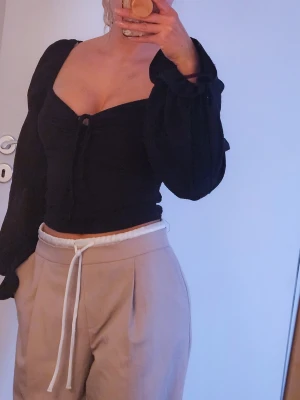 Svart blus med snörning - Trendig svart blus med långa puffärmar och snygg snörning framtill. Toppen har en fyrkantig ringning och är gjord i ett lätt, krinklat tyg som ger en cool struktur. Bra stretch pga smycken och använd en eller två ggr.