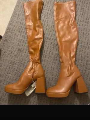 Bruna knähöga boots med klack - Säljer ett par helt nya bruna knähöga boots från Zara med chunky blockklack och rundad tå. Stövlarna har dragkedja på insidan och är tillverkade i ett glansigt konstläder. Perfekta för att lyfta vilken outfit som helst.