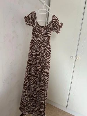 Brun långklänning med zebramönster - Säljer en brun långklänning med bash zebramönster och korta puffärmar. Klänningen har markerad midja och lätt rynkad byst, samt offshoulder-skärning. Perfekt för dig som vill sticka ut med ett djurmönstrat plagg! Köpte den och den passade tyvärr inte på mig😊
