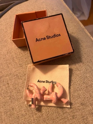 Rosa örhängen från Acne Studios - Unika örhängen från Acne Studios i en ljusrosa färg med vriden, chunky design. Tillverkade i ett glansigt material som ger en cool och modern look. Kommer med original ask(som tyvärr blivit skadad av vatten) och tygpåse från Acne Studios. Använda ca 3 gånger, som nya.