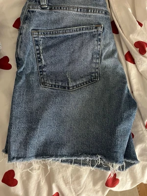 Blå jeansshorts med rå kant - Säljer ett par klassiska blå jeansshorts från 157 med låg midja och rå, fransig kant nedtill. Shortsen har fem fickor, knappgylf och en avslappnad passform. Perfekta för sommardagar och enkel att matcha med olika toppar.