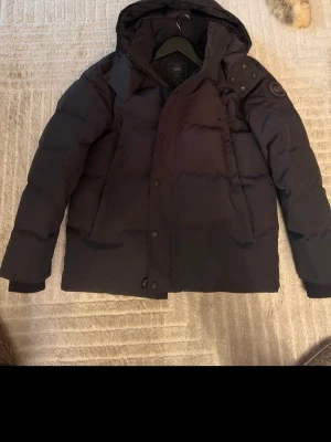 Svart dunjacka från Canada Goose - Säljer en svart dunjacka från Canada Goose med huva och diskret logga på ärmen. Jackan har dragkedja och knappar framtill samt två stora fickor. Perfekt för kalla vinterdagar och riktigt varm tack vare dunfyllningen. Storlek M. Ingen kopia, äkta jacka så inga skambud!