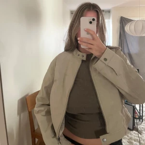 Beige skinnjacka med dragkedja - Säljer en beige skinnjacka med rak passform och snygga detaljer som ståkrage med tryckknapp och dragkedja framtill. Jackan har långa ärmar och minimalistisk design som ger en clean look. Nyinköpt för några månader sen men knappt använd så i nyskick! Nypris var 799kr.