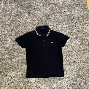 Svart Fred Perry pikétröja  - Klassisk svart pikétröja från Fred Perry i 100% bomull. Tröjan har vit broderad logga på bröstet, vitrandiga detaljer på krage och ärmslut samt knäppning framtill. Perfekt för en stilren och sportig look.