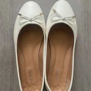 Vita ballerinaskor med rosett - Snygga vita ballerinaskor från DAY VINE med rund tå och dekorativ rosett framtill med gulddetaljer. Skorna har en klassisk och stilren design, perfekt till jeans eller klänning. 