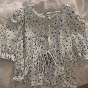 Blommig blus med knyt  - Supersöt blommig blus från Aware i vitt med små blå blommor. Blusen har v-ringning, knyt framtill och volangdetaljer vid ärmslut och midja. Lätt och luftig med korta ärmar, perfekt för varma dagar.