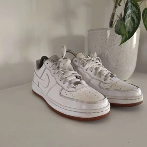 Nike Air Force 1 vita sneakers - Klassiska Nike Air Force 1 sneakers i vitt skinn med vit ovandel, perforerade detaljer och snörning. Skorna har en brun gummisula som ger en snygg kontrast. Perfekt för dig som gillar en clean och tidlös look.