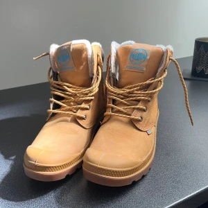 Palladium vinterboots i mocka, strl 41 - Snygga ljusbruna vinterboots från Palladium i mocka med grov sula och snörning. Fodrade med mjuk, vit syntetpäls för extra värme. Vattentäta och perfekta för kalla dagar. Märke och detaljer i blått på plös och häl. Klassisk outdoor-look.