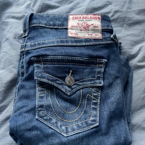 True Religion Joey jeans blå denim - Snygga blå jeans från True Religion, modell Joey. Klassisk femficksdesign med markerade kontrastsömmar och stora ficklock bak med logoknappar. Låg midja och bootcut-silhuett. Tillverkade i mjuk denim med coola detaljer. Det är i storlek w27 och passar mig perfekt som brukar ha S och är 173cm lång!