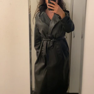 Svart skinn trenchcoat med bälte - Säljer en svart trenchcoat i skinnimitation med bälte i midjan och stora slag. Jackan har långa ärmar, knappar framtill och en snygg, oversized passform. Perfekt för dig som vill ha en stilren och edgy look. 