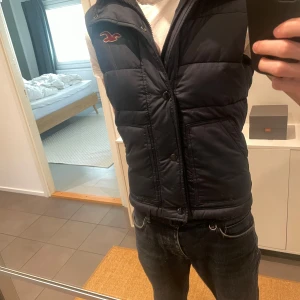 Mörkblå dunväst från Hollister - Snygg mörkblå dunväst från Hollister med hög krage, dragkedja och knappar framtill. Västen har två stora fickor och Hollisters logga broderad på bröstet. Perfekt för lager-på-lager och chill höst- och vinterstil.