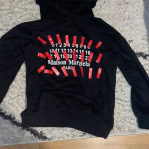 Maison Margiela svart hoodie med tryck - Svart hoodie från Maison Margiela med ikoniskt vitt och rött tryck på magen. Riktigt fet nu till vintern i bra skick och bra pris!