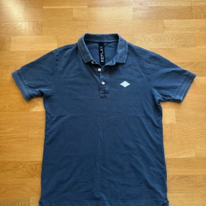 Blå pikétröja från Replay - Snygg marinblå pikétröja från Replay med klassisk krage, knappar framtill och logga på bröstet. Kragen har Replay-tryck på baksidan för en extra cool detalj. Perfekt till jeans eller shorts.