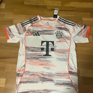 Bayern München bortatröja 25/26 - Snygg FC Bayern München fotbollströja från Adidas i vitt med grafiskt mönster i rött och grått. Tröjan har korta ärmar, orange detaljer och klubbmärke samt sponsorer tryckta på bröst och ärm. Tillverkad i lätt och ventilerande material (AEROREADY).