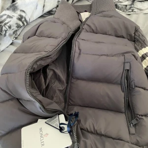 Grå dunväst från Moncler - Snygg grå dunväst från Moncler med quiltad design och ribbad krage. Västen har dragkedja framtill och två praktiska fickor med dragkedja. Perfekt för lager-på-lager och har en klassisk Moncler-logga på bröstet.