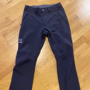 Svarta täckbyxor från Haglöfs - Svarta täckbyxor från Haglöfs med dragkedjeförsedda fickor och mjukt fleecefoder på insidan. Byxorna har bälteshällor, knapp och dragkedja i midjan samt diskret Haglöfs-logga på benet. Perfekta för kalla dagar utomhus.