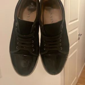 Svarta sneakers från Lanvin - Snygga svarta sneakers från Lanvin med klassisk rund tå och snörning. Ovandelen är i mocka med detaljer i blankt skinn på tån och runt sulan. Skorna har vit sula och diskret logga på innersulan. Perfekta för dig som gillar stilrena och lyxiga sneakers.