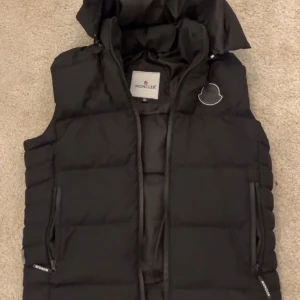 Svart dunväst från Moncler - Säljer en svart dunväst från Moncler med klassisk logga på bröstet och dragkedjor på fickorna. Västen har en hög krage och quiltad design som ger en clean och modern look. Perfekt att slänga över en hoodie eller tröja när det är kyligt ute.
