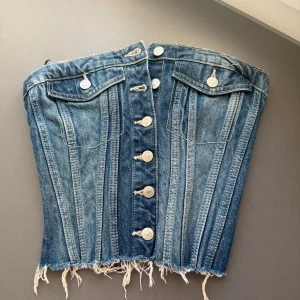 Jeanskorsett från Zara med knappar - Säljer en snygg jeanskorsett från Zara i blå denim med silverfärgade knappar framtill, två fejkfickor och rå fransad nederkant. Toppen är axelbandslös och har smock baktill för extra komfort. Perfekt för dig som vill sticka ut med en cool och trendig look.