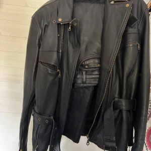 Svart skinnställ  - Svart skinnjacka med flera dragkedjor, bälte i midjan och detaljer i metall.                                Svarta skinnbyxor med midjemått 54 och innerbenslängd 78. Det är i nyskick. 