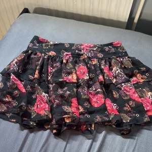 Blommig kortkjol från Divided - Supersöt kortkjol från Divided med svart botten och stora blommor i rosa, lila och orange toner. Kjolen har volanger, små metallöljetter och ett luftigt, lätt tyg som ger en flowy känsla. Perfekt för dig som vill sticka ut med en färgglad och lekfull stil. Passar även bra med tex en vit kofta.