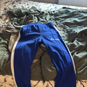 Blåa Palm Angels mjukisbyxor - Snygga blåa mjukisbyxor från Palm Angels med vita sidoränder och broderad logga på låret. Byxorna har elastisk midja och raka ben, perfekta för en chill och sportig look. Materialet är mjukt och följsamt, vilket gör dem riktigt bekväma.