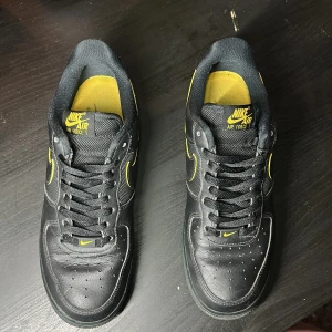 Nike Air Force 1 svart/gul sneakers - Svarta Nike Air Force 1 sneakers med gula detaljer och logga. Skorna har klassisk rund tå, snörning och platt sula. Ovandelen är i en mix av skinn och mesh för extra komfort och stil. Perfekta för dig som vill sticka ut med en sportig vibe.