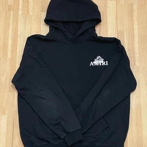 Svart hoodie från Amiri med ängeltryck - Svart hoodie från Amiri i 100% bomull med vit Amiri-logga och liten ängel på bröstet samt stort ängeltryck och logga på ryggen. Klassisk passform med huva och känguruficka. Perfekt för dig som gillar streetwear med exklusiv vibe. Priset kan alltid diskuteras