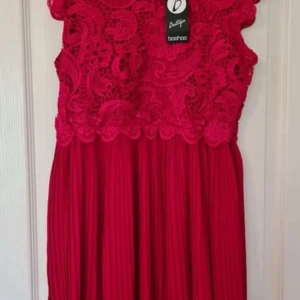 Ny Röd spetsklänning från Boohoo - Säljer en röd klänning från Boohoo med vacker spets upptill och plisserad kjol. Klänningen har ärmlös design och detaljerad spets som ger en lyxig känsla. Perfekt för dig som vill sticka ut med färg och snygga detaljer.