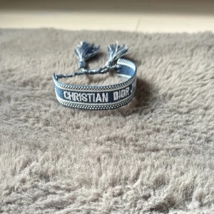 Blå Christian Dior armband med tofsar - Snyggt blått armband från Christian Dior med vit broderad text 'CHRISTIAN DIOR J'ADIOR'. Armbandet har en vävd design med små vita detaljer och avslutas med två coola tofsar i matchande blå nyans. Perfekt accessoar för att lyfta din outfit.