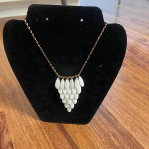 Guldigt halsband med vita droppar - Unikt halsband med guldtonad kedja och hängande vita droppformade pärlor i flera lager. Kedjan är tunn och smidig med karbinlås. Perfekt för dig som vill sticka ut med ett snyggt statement-smycke.