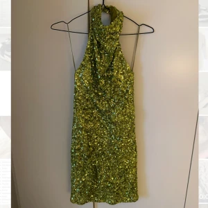 Grön paljettklänning med öppen rygg - Snygg grön kort klänning täckt av glittrande paljetter. Hög halsringning och öppen rygg.Perfekt för dig som vill sticka ut och glänsa. Klänningen har en figurnära passform och är fodrad på insidan för extra komfort. Använd vid ett tillfälle, så den är i riktigt bra skick! 