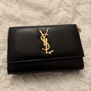 Svart axelväska i skinn - Snygg svart axelväska med klassisk guldlogga framtill och guldfärgad kedja. Väskan är tillverkad i slätt skinn och har en stilren, rektangulär form som ger en lyxig känsla. Perfekt för dig som vill ha en ikonisk accessoar med exklusiva detaljer. Den är köpt utomlands, av ngt random märke, inget viktigt märke.